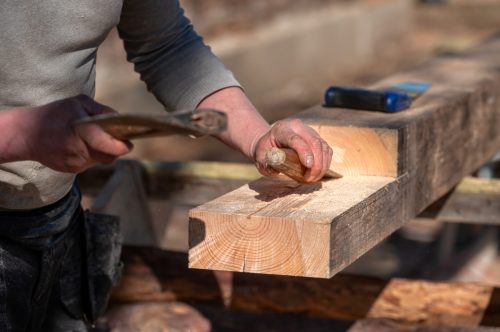 A carpenter skillfully shapes a wooden beam using a chisel and hammer in sunlight.